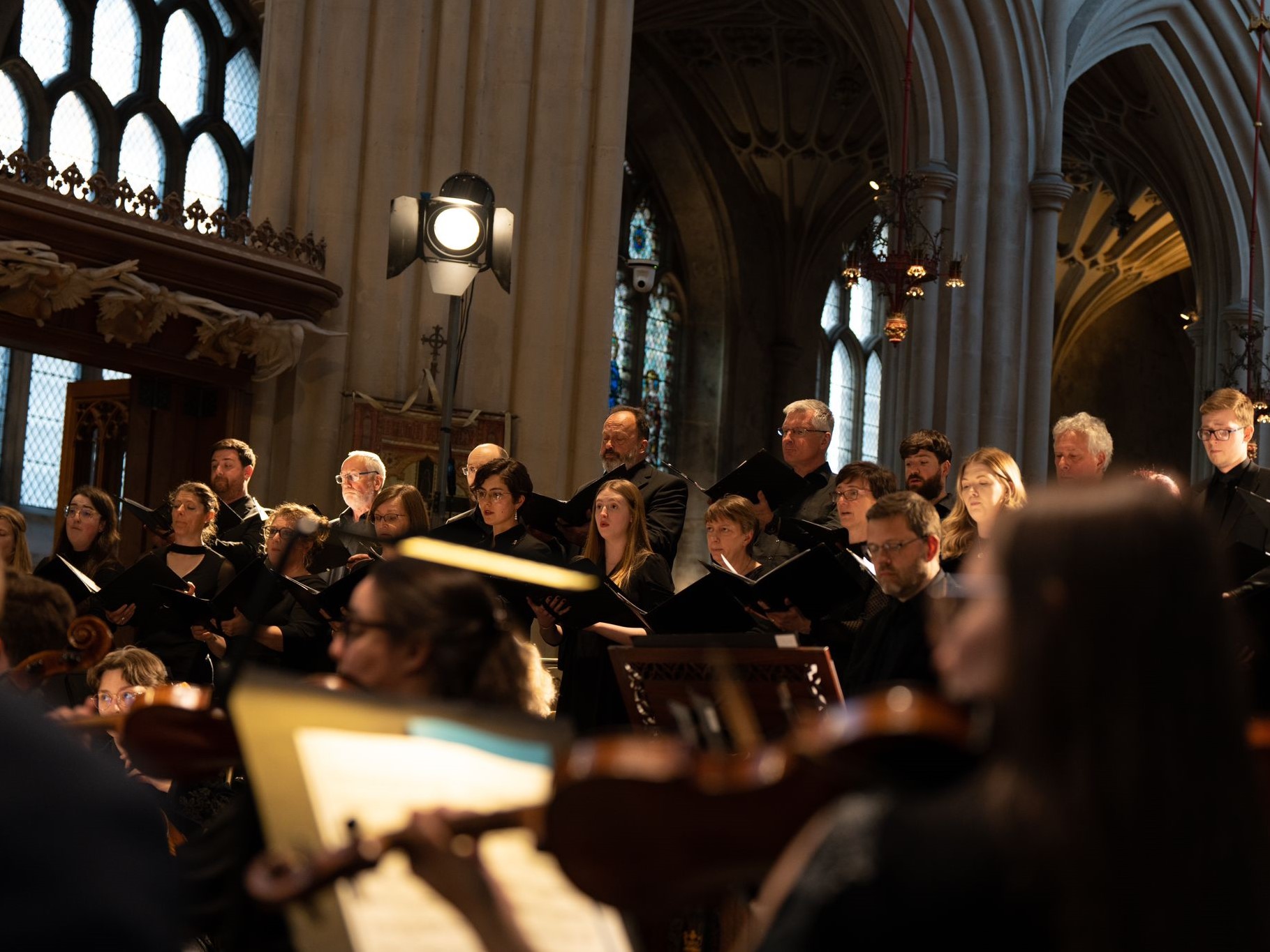 A colour photograph of Bath Festival Orchestra and Bath Camerata performing Mozart's Requiem in Bath Abbey. The choir is behind the orchestra and some of the church architecture is in view